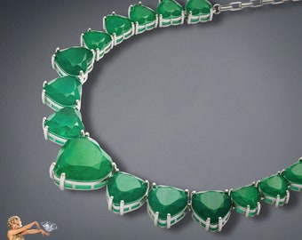 Op de afbeelding: Een luxe ketting met grote, driehoekige, smaragdgroene edelstenen gezet in een zilverkleurig metaal. De ketting wordt gepresenteerd tegen een donkergrijze achtergrond, die de levendige groene kleur van de edelstenen benadrukt.