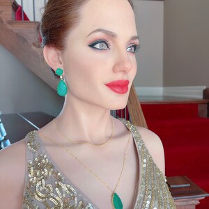 May include: A woman wearing a gold sequined dress, gold necklace, and emerald green earrings. The earrings are a teardrop shape. The woman has red lipstick and dark eye makeup. The background includes a staircase and red carpet.