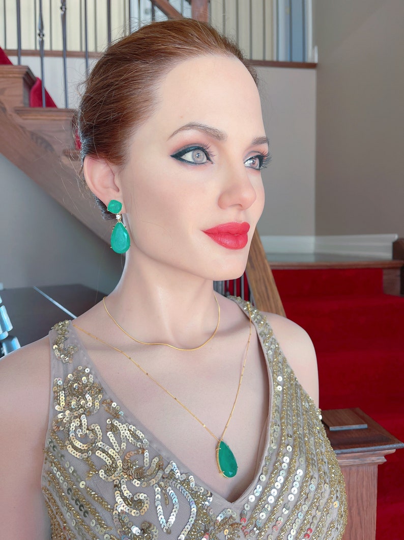 May include: A woman wearing a gold sequined dress, gold necklace, and emerald green earrings. The earrings are a teardrop shape. The woman has red lipstick and dark eye makeup. The background includes a staircase and red carpet.