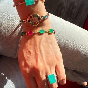 May include: A collection of gold-toned jewelry, including a bracelet with a large emerald-cut green gemstone, a watch with a rainbow-colored bezel, and a ring with a matching green gemstone. The jewelry is worn on a wrist and hand.