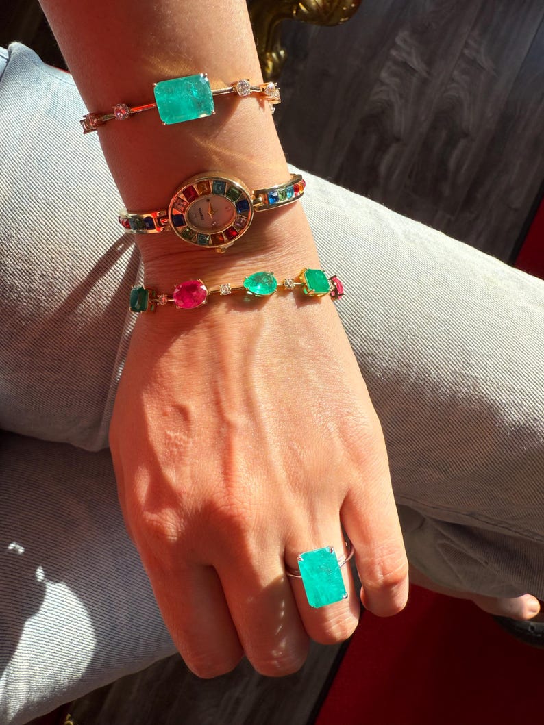 May include: A collection of gold-toned jewelry, including a bracelet with a large emerald-cut green gemstone, a watch with a rainbow-colored bezel, and a ring with a matching green gemstone. The jewelry is worn on a wrist and hand.