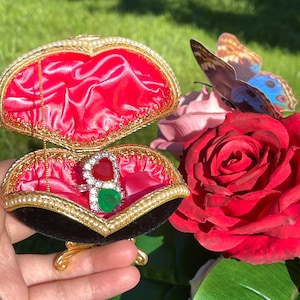 May include: A red, green, and white jeweled ring sits inside a black velvet heart-shaped ring box with gold trim. The box is open and rests on a gold stand. The box is surrounded by flowers.