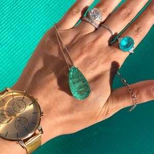 May include: A gold wristwatch with a gold mesh band and a gold face. A silver necklace with a large, teardrop-shaped, green gemstone pendant. Several rings with clear and blue gemstones.