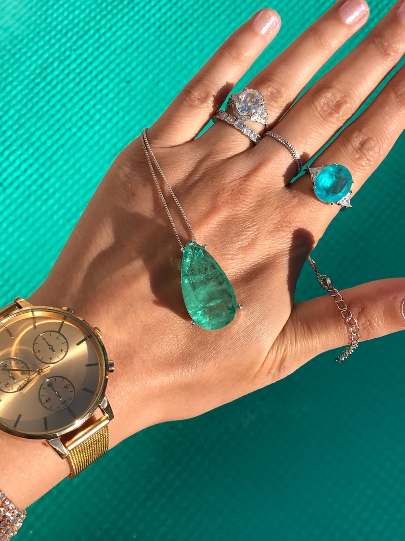 May include: A gold wristwatch with a gold mesh band and a gold face. A silver necklace with a large, teardrop-shaped, green gemstone pendant. Several rings with clear and blue gemstones.