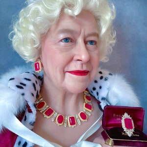 May include: A woman wearing a crown and a fur-trimmed robe holds a red velvet box with a gold and red jeweled brooch inside. The box has the words "Bulgari" and "Made in America" on it.