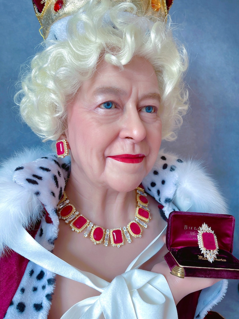 May include: A woman wearing a crown and a fur-trimmed robe holds a red velvet box with a gold and red jeweled brooch inside. The box has the words "Bulgari" and "Made in America" on it.