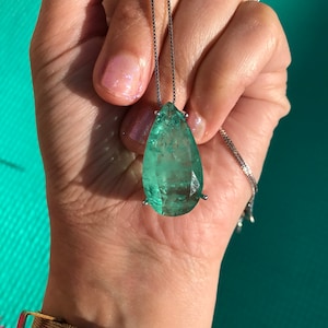 May include: A teardrop-shaped, emerald-green gemstone pendant necklace. The large, faceted stone is suspended from a delicate silver chain. The pendant is held in place by a silver setting. The background is a turquoise color.
