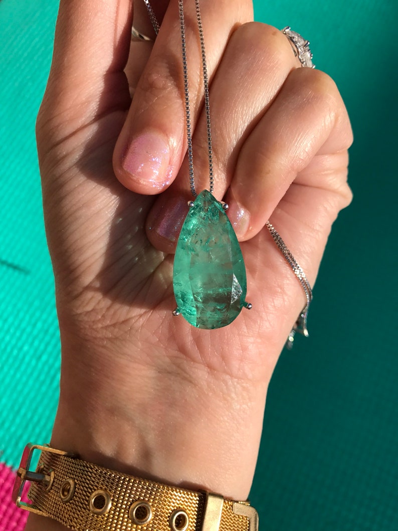May include: A teardrop-shaped, emerald-green gemstone pendant necklace. The large, faceted stone is suspended from a delicate silver chain. The pendant is held in place by a silver setting. The background is a turquoise color.