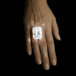 May include: A large, rectangular-cut diamond ring. The diamond is set on a hand against a black background. The diamond sparkles brightly, reflecting light.
