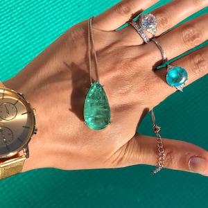 May include: A collection of jewelry including a silver necklace with a large, pear-shaped, green gemstone pendant. Also visible are several rings with large gemstones and a gold wristwatch with a mesh band.