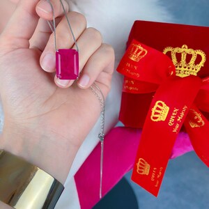 May include: A silver chain necklace with a large, rectangular, pink gemstone pendant. The pendant is set in a silver frame. The necklace is being held by a hand, and a red velvet box with gold crowns and the text "Queen Your Majesty" is in the background.