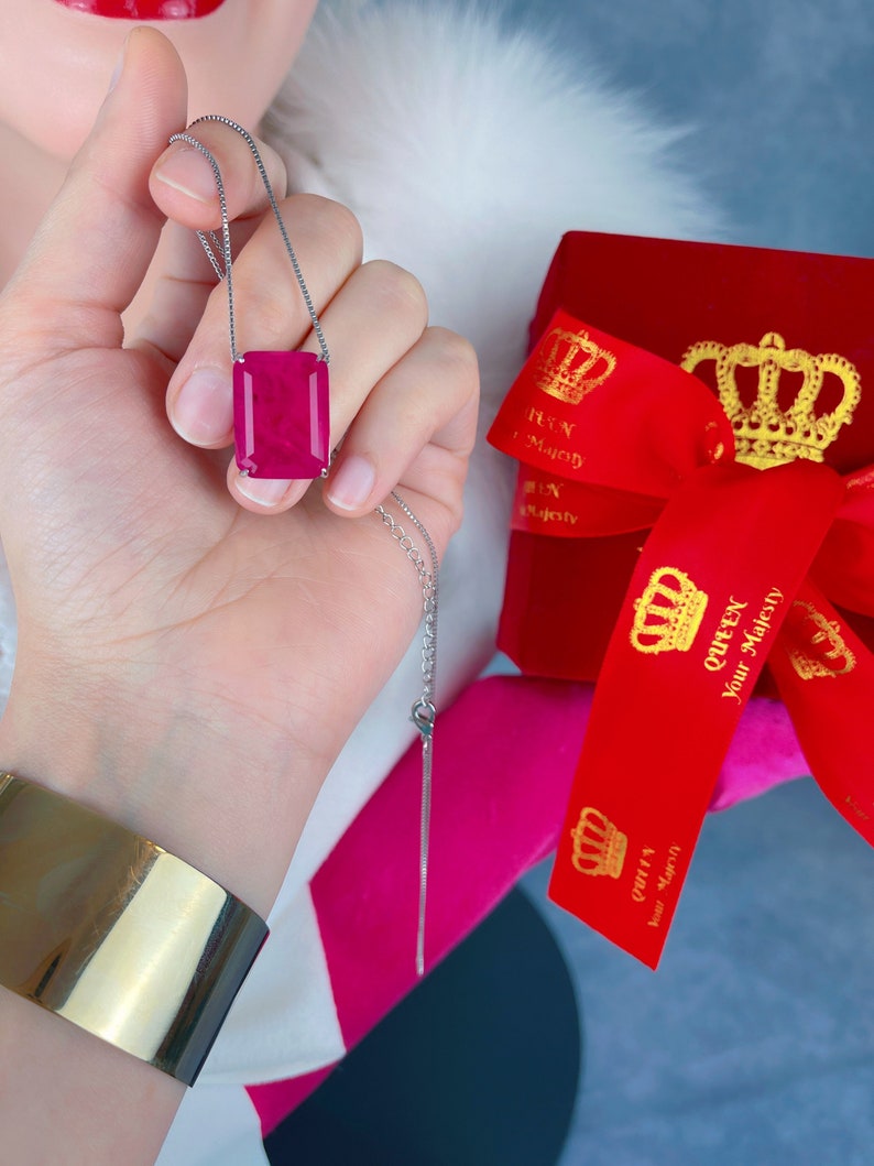 May include: A silver chain necklace with a large, rectangular, pink gemstone pendant. The pendant is set in a silver frame. The necklace is being held by a hand, and a red velvet box with gold crowns and the text "Queen Your Majesty" is in the background.