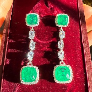 May include: A pair of emerald green and diamond dangle earrings. The earrings feature a square emerald green stone surrounded by smaller diamonds, followed by three smaller diamonds hanging below.