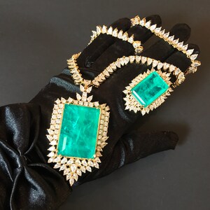 Op de afbeelding: Een gouden ketting en hanger set met grote smaragdgroene edelstenen omringd door heldere zirkonia stenen. De ketting en hanger liggen op een zwarte satijnen achtergrond.