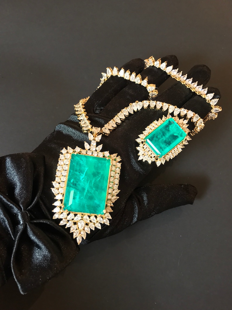 Op de afbeelding: Een gouden ketting en hanger set met grote smaragdgroene edelstenen omringd door heldere zirkonia stenen. De ketting en hanger liggen op een zwarte satijnen achtergrond.