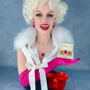 May include: A white mannequin bust with blonde hair wearing a white lace top, pink gloves, and a pink bow. The mannequin is holding a small white box with a red jewel inside. The box is labelled "Diamonds".