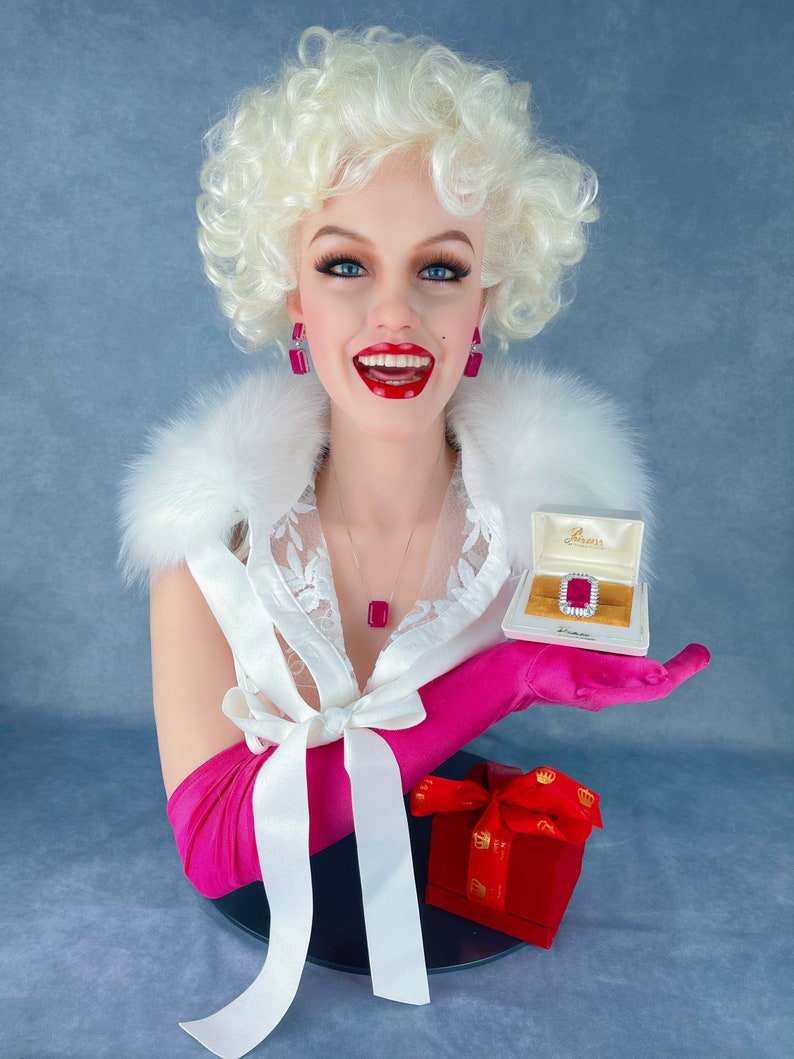 May include: A white mannequin bust with blonde hair wearing a white lace top, pink gloves, and a pink bow. The mannequin is holding a small white box with a red jewel inside. The box is labelled "Diamonds".