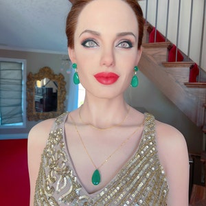 May include: A woman wearing a gold sequined dress, a gold necklace, and green gemstone earrings and pendant. The woman has red lipstick and is standing in front of a staircase with a red carpet.