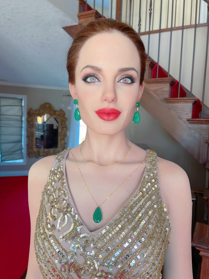 May include: A woman wearing a gold sequined dress, a gold necklace, and green gemstone earrings and pendant. The woman has red lipstick and is standing in front of a staircase with a red carpet.