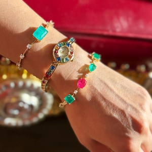 May include: A gold-toned watch and two bracelets are displayed on a wrist. The watch has a round face with a rainbow-colored bezel. One bracelet features a large rectangular turquoise stone, and the other has colorful gemstones.