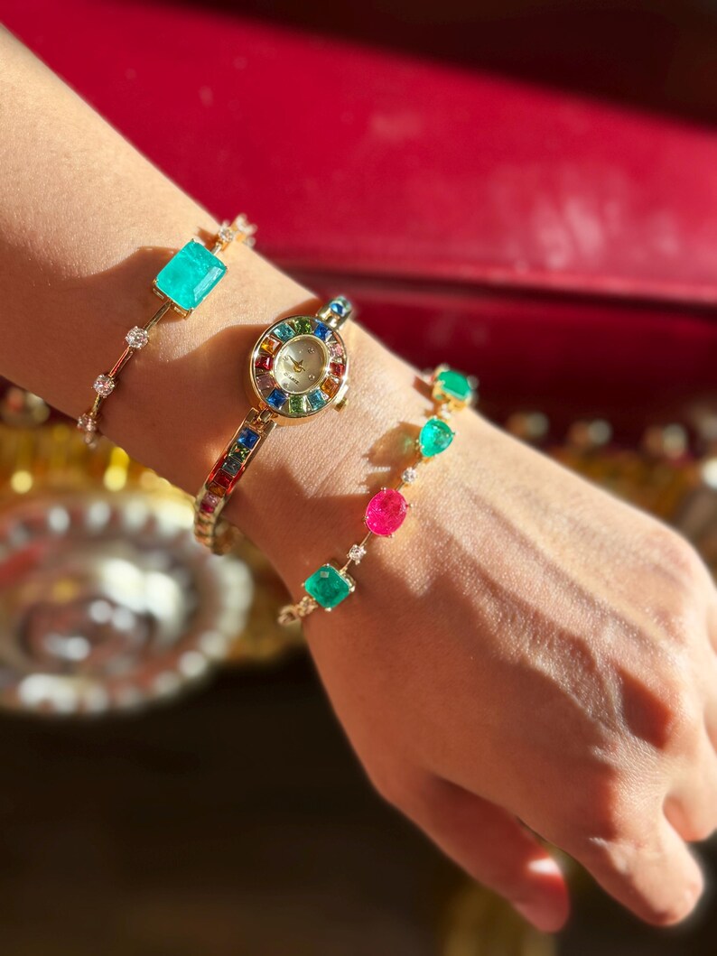 May include: A gold-toned watch and two bracelets are displayed on a wrist. The watch has a round face with a rainbow-colored bezel. One bracelet features a large rectangular turquoise stone, and the other has colorful gemstones.