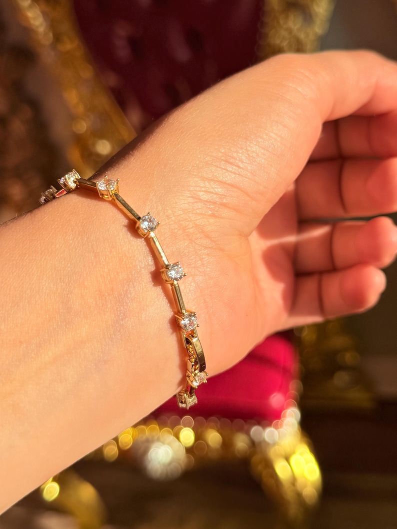 May include: A gold bracelet with multiple clear, round gemstones. The bracelet is made of gold-colored metal and is worn on a wrist. The gemstones are evenly spaced along the bracelet's length, reflecting light.