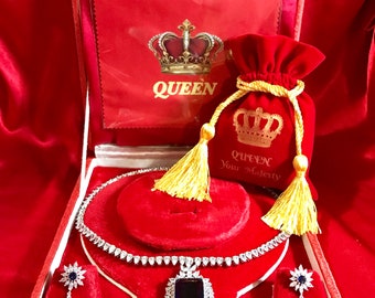 May include: A red velvet jewelry box with a gold crown and the word "QUEEN" on the lid. The box contains a silver necklace with a large blue stone pendant, a pair of matching earrings, and a small red velvet bag with a gold crown and the words "QUEEN Your Majesty".