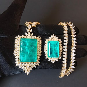 Op de afbeelding: Een gouden ketting met een grote hanger van smaragdgroene steen, omringd door kleinere heldere stenen. De ketting ligt op een zwart oppervlak.
