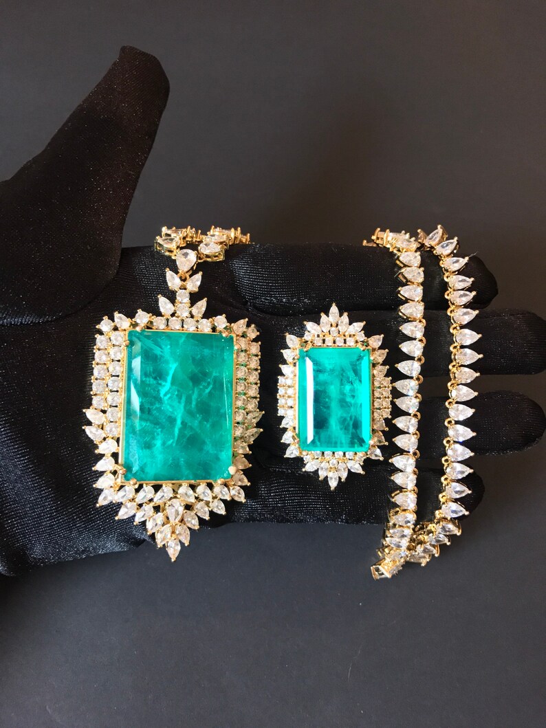 Op de afbeelding: Een gouden ketting met een grote hanger van smaragdgroene steen, omringd door kleinere heldere stenen. De ketting ligt op een zwart oppervlak.