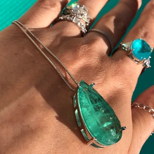 May include: A silver necklace with a large, teardrop-shaped, teal-colored pendant. The pendant is set in a silver frame. Several rings with large gemstones are also visible.