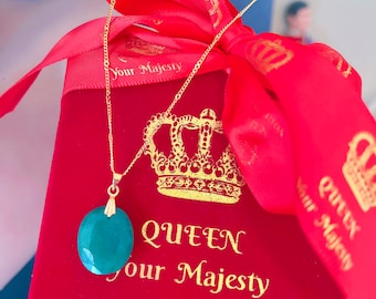 Könnte beinhalten: Eine goldene Halskette mit einem großen, ovalen, türkisfarbenen Anhänger. Die Halskette wird auf einer roten Schachtel mit goldenem Text "QUEEN Your Majesty" präsentiert. Ein rotes Band mit goldenem Text und einem Kronenmuster umgibt die Schachtel.
