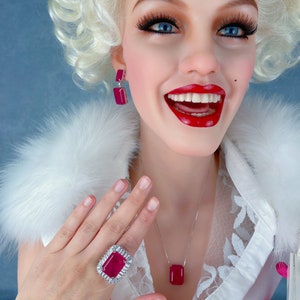 May include: A woman wearing a white blouse with a fur collar and a pink skirt. She is wearing a silver necklace with a large red gemstone pendant, a silver ring with a large red gemstone, and silver earrings with red gemstones.