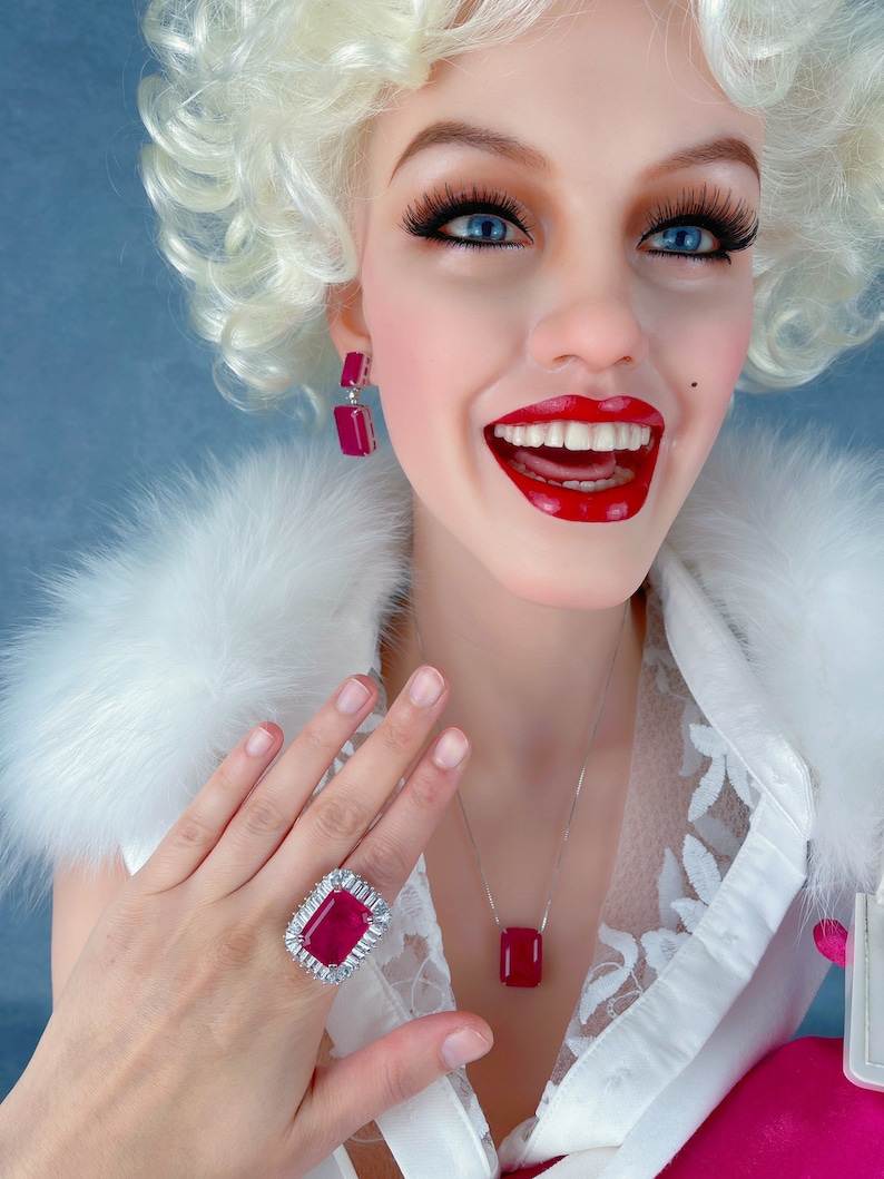 May include: A woman wearing a white blouse with a fur collar and a pink skirt. She is wearing a silver necklace with a large red gemstone pendant, a silver ring with a large red gemstone, and silver earrings with red gemstones.