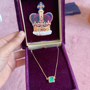 May include: A gold chain necklace with a square green gemstone pendant in a velvet-lined box. The box is decorated with a gold crown.