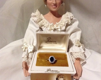 Könnte beinhalten: Puppe in weißem Brautkleid, Schleier und Diadem, die eine Ringbox mit einem Saphir- und Diamantring präsentiert. Eine Karte mit "Diana Princess of Wales" ist ebenfalls sichtbar.