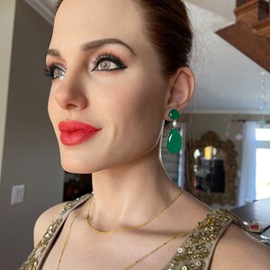 May include: A woman wearing gold-colored jewelry, including a necklace and earrings. The earrings are emerald green with a diamond accent. The woman is wearing a gold sequined dress. Her lips are red.