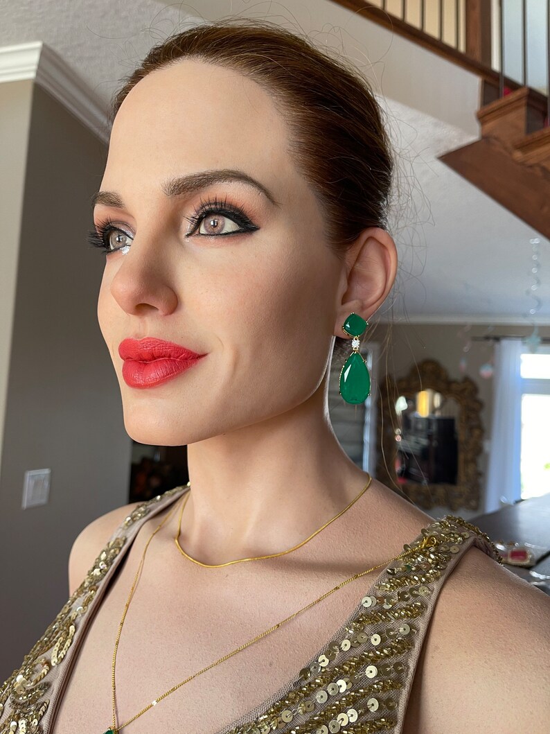 May include: A woman wearing gold-colored jewelry, including a necklace and earrings. The earrings are emerald green with a diamond accent. The woman is wearing a gold sequined dress. Her lips are red.