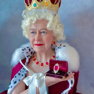May include: A woman wearing a red velvet robe with a white fur trim, a gold crown with red and green jewels, and a gold necklace with red jewels. She is holding a small red velvet box with a gold clasp and a red jewel inside. The box is labeled "BLOVA"