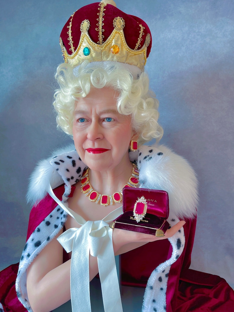 May include: A woman wearing a red velvet robe with a white fur trim, a gold crown with red and green jewels, and a gold necklace with red jewels. She is holding a small red velvet box with a gold clasp and a red jewel inside. The box is labeled "BLOVA"