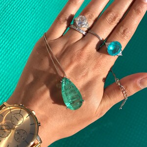 May include: A collection of jewelry including a gold watch, rings, and a pendant necklace. The rings feature large, clear and blue gemstones. The pendant has a large, teardrop-shaped, green gemstone. The watch has a gold band and face.