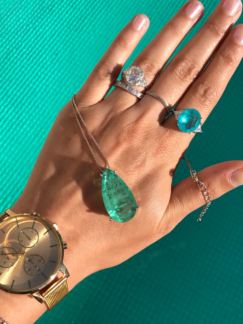 May include: A collection of jewelry including a gold watch, rings, and a pendant necklace. The rings feature large, clear and blue gemstones. The pendant has a large, teardrop-shaped, green gemstone. The watch has a gold band and face.