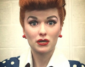 May include: A woman with red hair styled in a 1950s updo, wearing a blue and white polka dot dress with a white collar and a red beaded necklace. She is wearing red lipstick and red earrings.