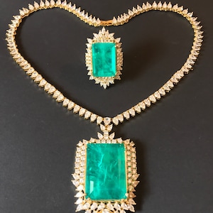 Op de afbeelding: Een gouden ketting met een grote hanger van smaragdgroene steen en een bijpassende ring. Beide stukken zijn bezet met kleinere, heldere stenen.