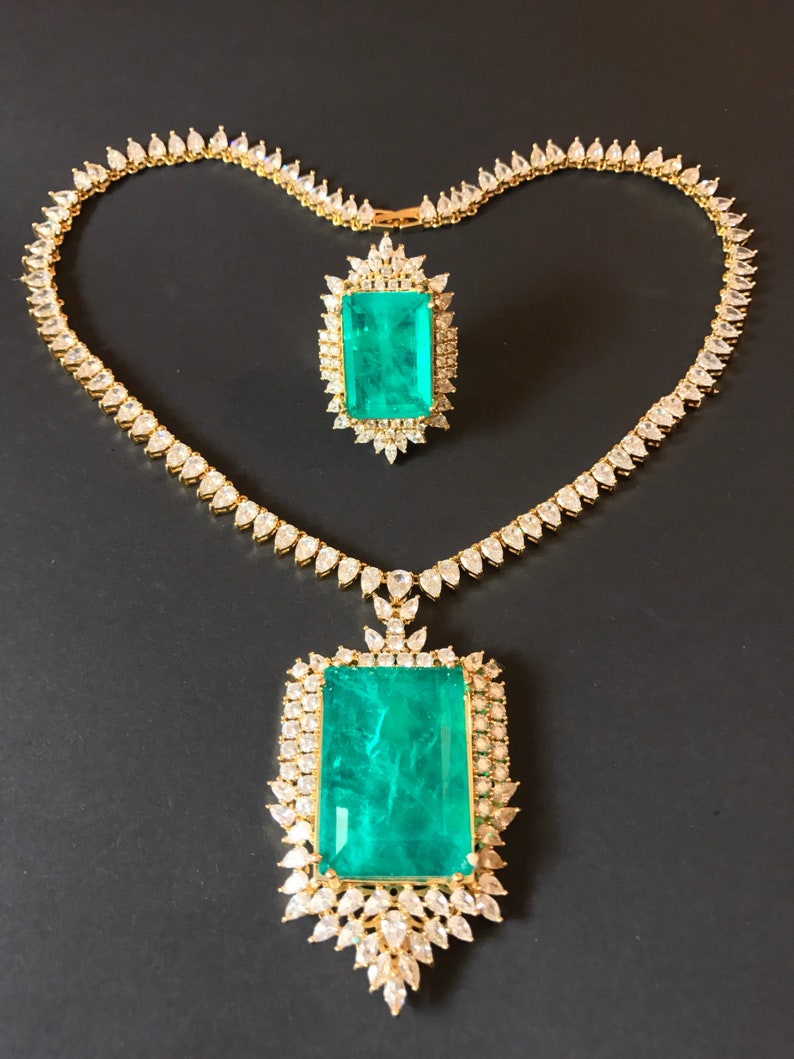 Op de afbeelding: Een gouden ketting met een grote hanger van smaragdgroene steen en een bijpassende ring. Beide stukken zijn bezet met kleinere, heldere stenen.