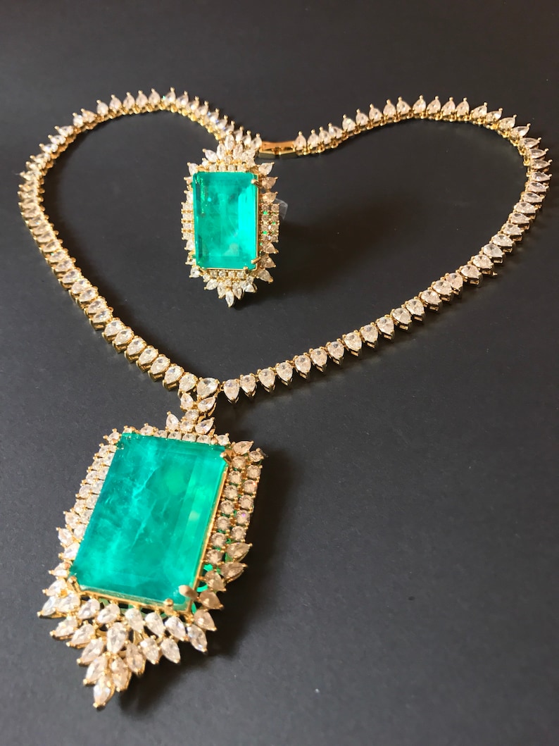 Op de afbeelding: Een gouden ketting- en ringset met grote smaragdgroene edelstenen omringd door kleinere heldere edelstenen.
