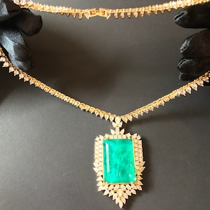 Op de afbeelding: Een gouden ketting met een grote hanger van smaragdgroene edelsteen, omringd door kleinere diamanten. De ketting is gemaakt van een delicate ketting met kleine diamant accenten.