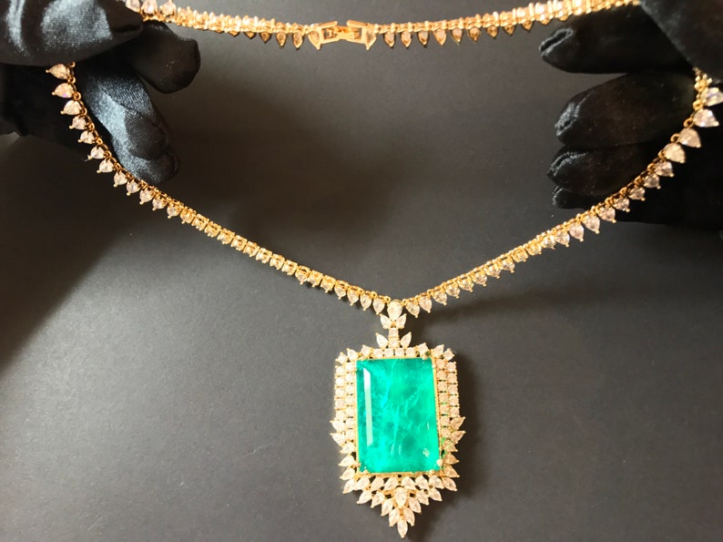 Op de afbeelding: Een gouden ketting met een grote hanger van smaragdgroene edelsteen, omringd door kleinere diamanten. De ketting is gemaakt van een delicate ketting met kleine diamant accenten.