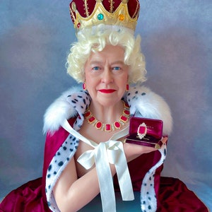 Puede incluir: Una figura de cera de una mujer que lleva una túnica de terciopelo rojo y armiño con una corona dorada y un collar de joyas rojas. Está sosteniendo una pequeña caja de terciopelo rojo con un anillo de oro en su interior.