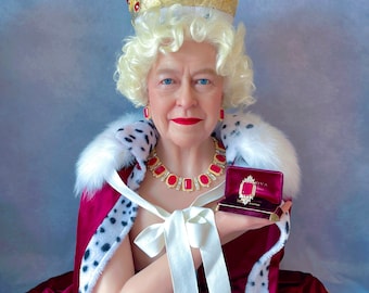 May include: A wax figure of a woman wearing a red velvet and ermine robe with a gold crown and a red jewel necklace. She is holding a small red velvet box with a gold ring inside.
