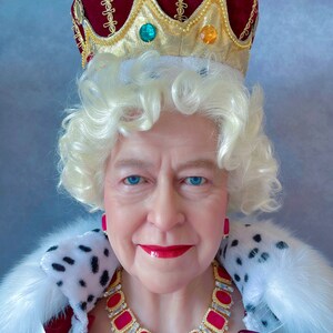 May include: A woman wearing a red velvet crown with gold trim and jewels, a white fur collar, and a red and gold necklace with large pink gemstones. She is wearing a white wig and has a serious expression.